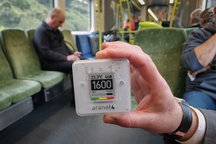 Hand holding a CO2 meter displaying 1600ppm, inside a train carriage