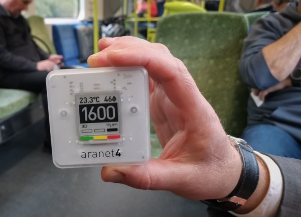 Hand holding a CO2 meter displaying 1600ppm, inside a train carriage