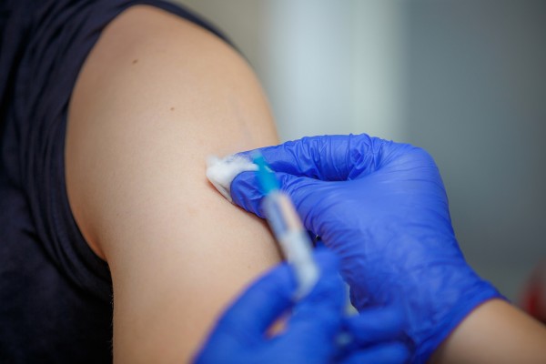 photograph of immunisation. A gloved hand holds a cotton swab to arm, syringe is in blurred foreground